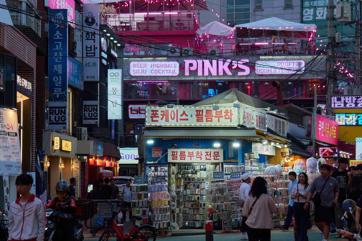 A bar street in Hongdae, Seoul at night with a neon sign reading BEER HIGHBALL SOJU COCKTAIL above a busy pedestrian street — soju highball global